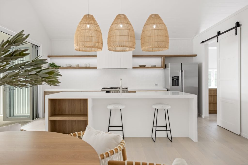 Inviting kitchen view highlighting rattan pendant lighting above a white island, complemented by wooden details and a fresh plant, with an open sliding door to the outdoor area.