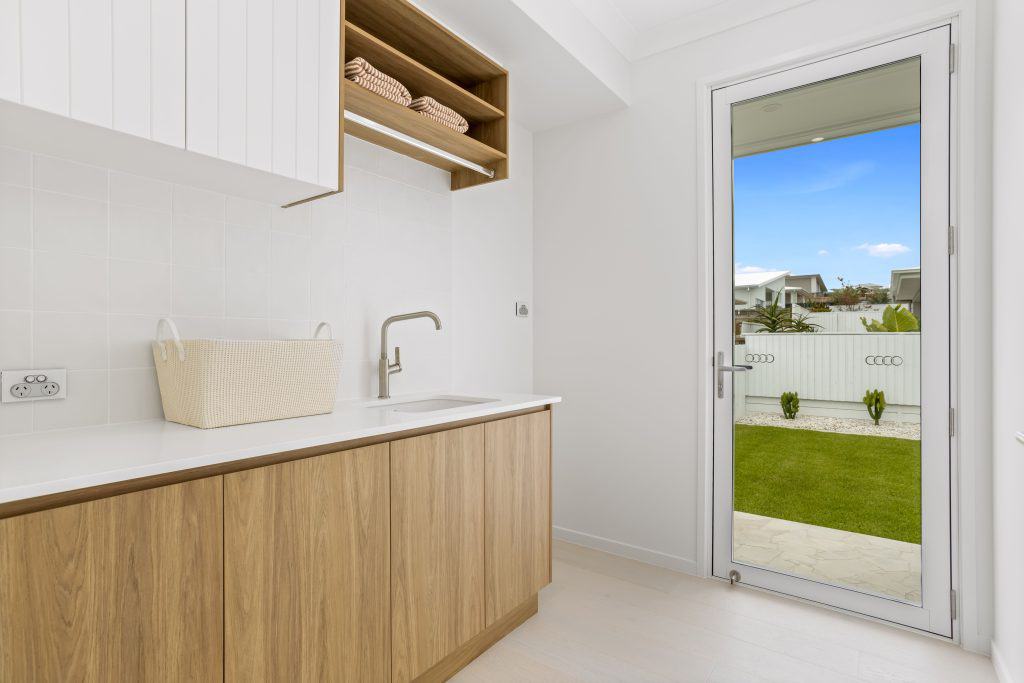 Laundry room with a crisp white countertop and wooden cabinetry, featuring a wicker basket and brushed nickel faucet, with a view to a well-kept garden through a glass door.