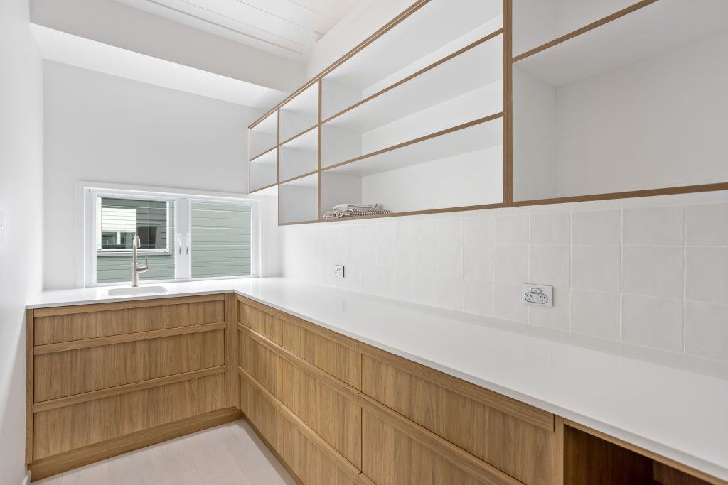 Laundry room featuring clean lines with white countertops, wooden cabinetry, and open shelving against a crisp white tiled wall, with a window offering natural light.