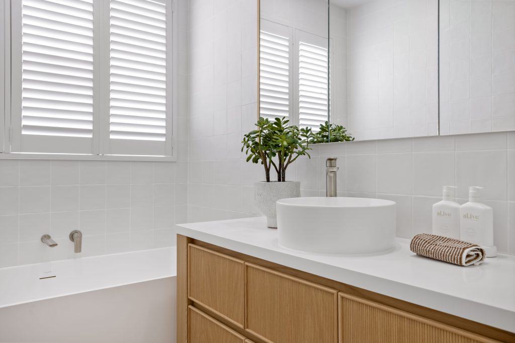 Minimalist bathroom vanity with a vessel sink, wooden cabinets, and white tile backdrop, complemented by natural light from the window and a green potted plant for a touch of freshness.