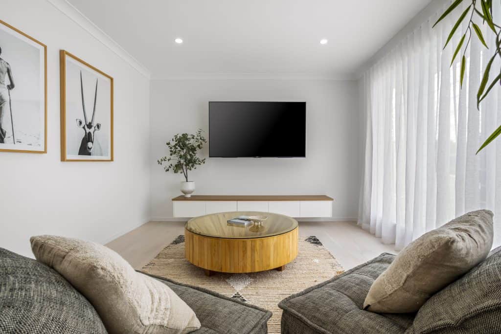 Cozy living room with soft grey sofas, a round wooden coffee table, and wall-mounted TV, accented by framed artwork and sheer curtains for a light, airy feel.