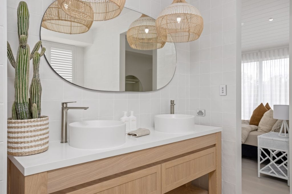 Chic bathroom vanity with twin vessel sinks, rattan pendant lights, and a large oval mirror, accented by a tall cactus in a woven planter, creating a natural, textured look.