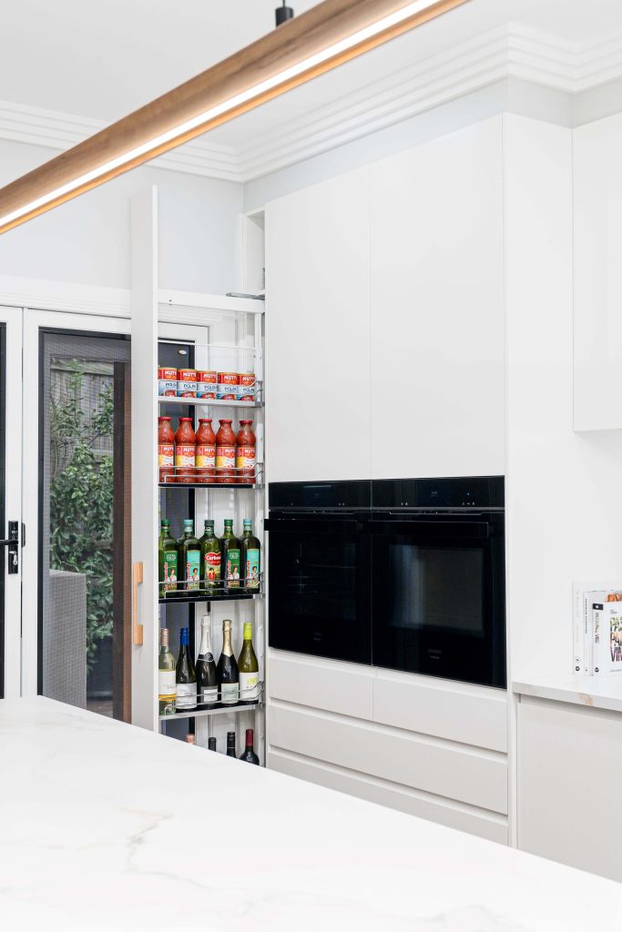 Contemporary white kitchen in Brisbane featuring a sleek pull-out pantry with organized shelves for oils, sauces, and wine bottles. The design includes integrated black double ovens and minimalist cabinetry, enhancing the clean and modern aesthetic.