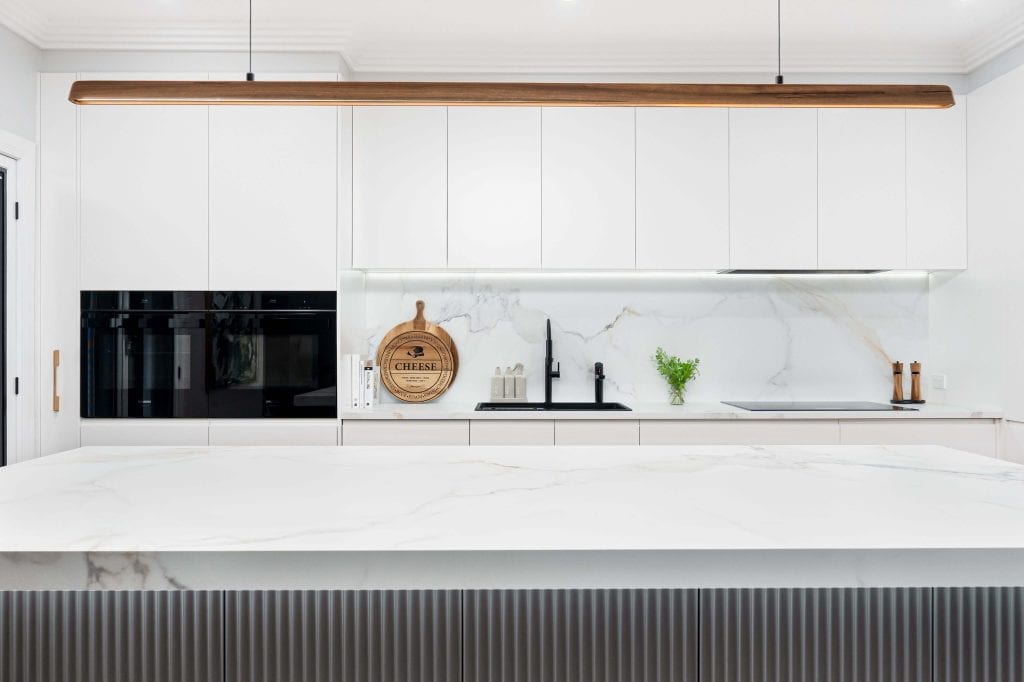 Modern white kitchen in Brisbane featuring a large marble island with ribbed detailing, black integrated appliances, and a sleek black matte faucet. The design includes a marble backsplash, minimalist white cabinetry, and wooden accents like a hanging light fixture and a decorative cheese board, creating a clean and elegant aesthetic.