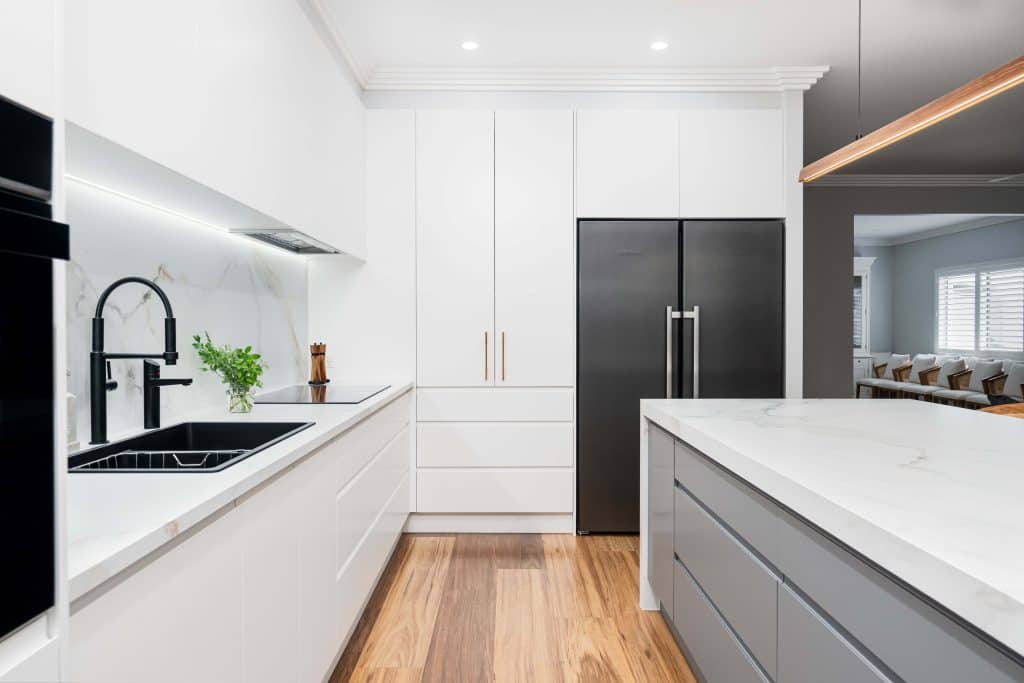 Contemporary white kitchen in Brisbane featuring a marble backsplash, black matte faucet, and integrated black appliances. The design includes sleek white cabinetry, a large kitchen island with a marble countertop, and wooden flooring. The minimalist aesthetic is enhanced by subtle wood and black accents, creating a clean and modern look.