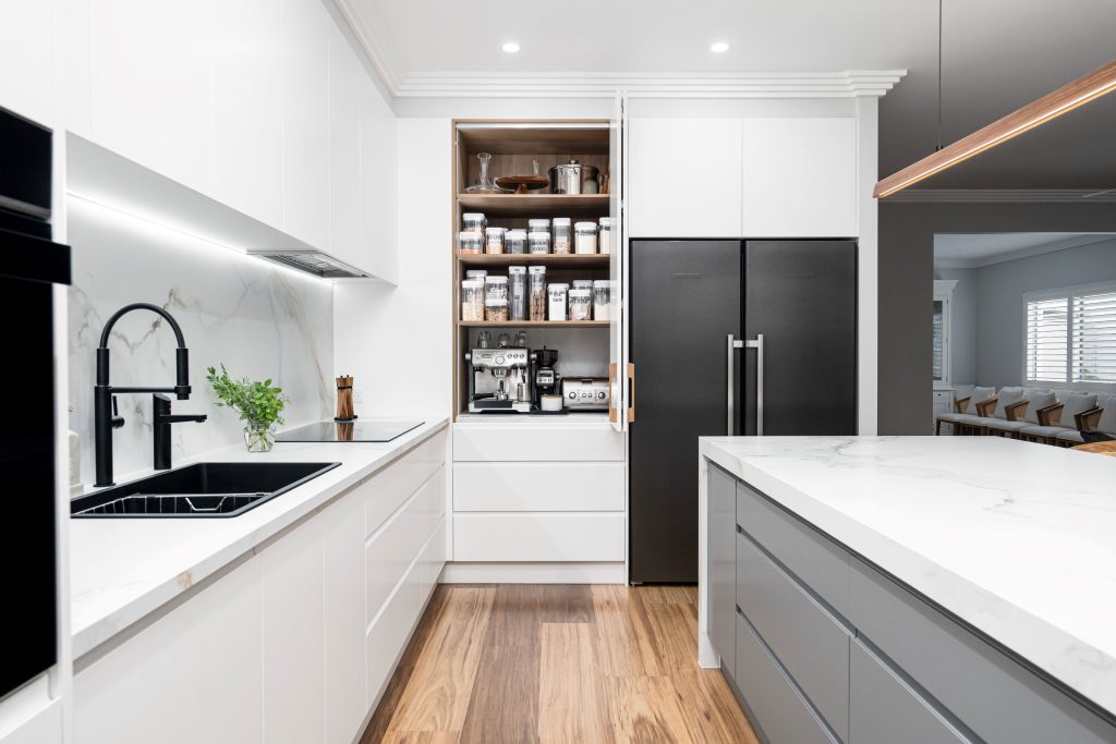 Modern white kitchen in Brisbane featuring a hidden coffee station with organized shelves for kitchen essentials and a built-in espresso machine. The design includes a marble backsplash, black matte faucet, integrated black appliances, and a large kitchen island with a marble countertop. The space is bright and functional, with a sleek and minimalist aesthetic.
