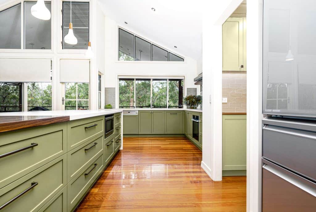 Modern kitchen renovation in Brisbane featuring olive green cabinetry with sleek black handles, polished wooden floors, and large windows offering natural light and forest views. The spacious design includes a central island with a white countertop and integrated appliances.