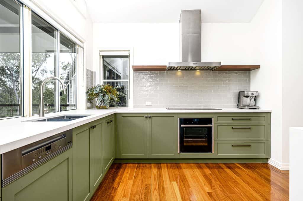 Stylish kitchen remodel in Brisbane featuring olive green cabinets, a sleek stainless steel range hood, and a modern electric stove. The design includes white countertops, a large sink under wide windows for ample natural light, and a glossy subway tile backsplash. The kitchen also boasts polished hardwood floors and contemporary kitchen appliances.