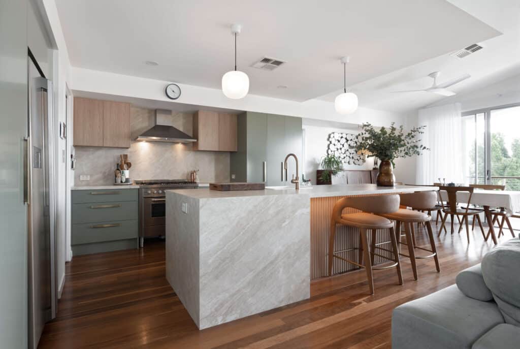 Modern open-plan kitchen with a natural stone waterfall benchtop, matte green cabinetry with brass handles, and upper cabinets in a light woodgrain finish. The design features integrated appliances, warm timber flooring, and a spacious island with a fluted timber panel and brushed brass tapware.