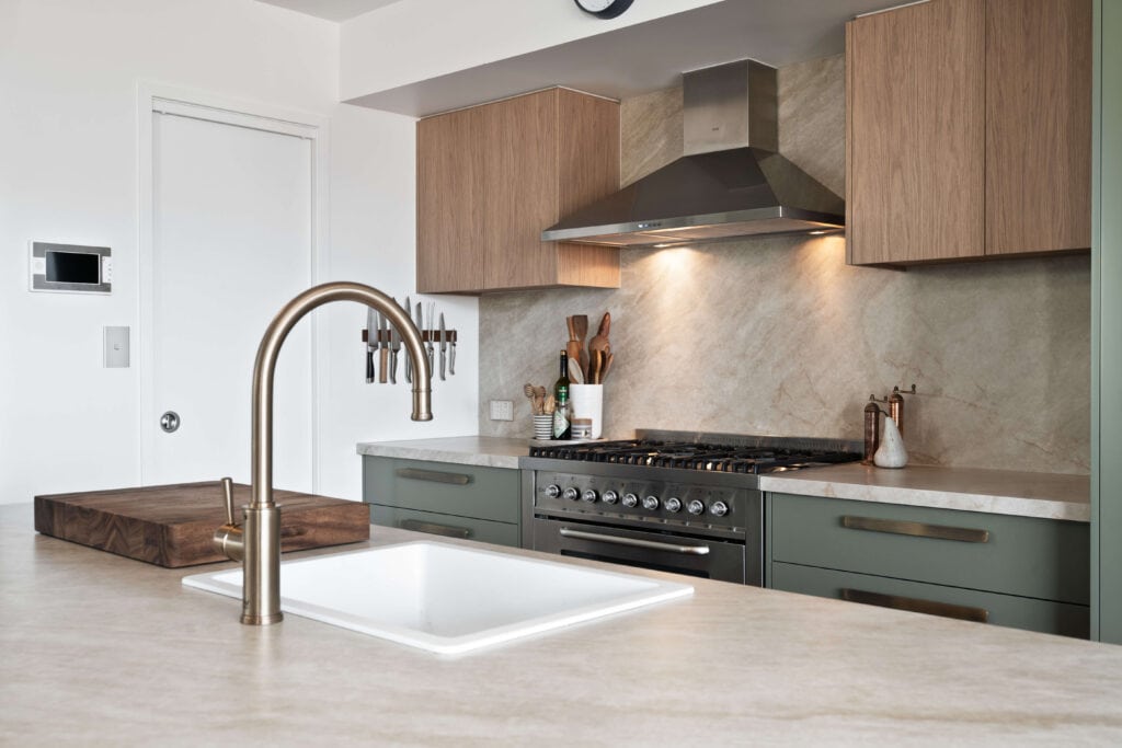 Detail of a modern kitchen with a natural stone benchtop and brushed brass tapware, complemented by sage green lower cabinetry with brass handles and warm woodgrain upper cabinets. The design includes a built-in stainless steel gas range with a matching rangehood, an integrated knife rack, and a large timber chopping board, set against a textured stone splashback.