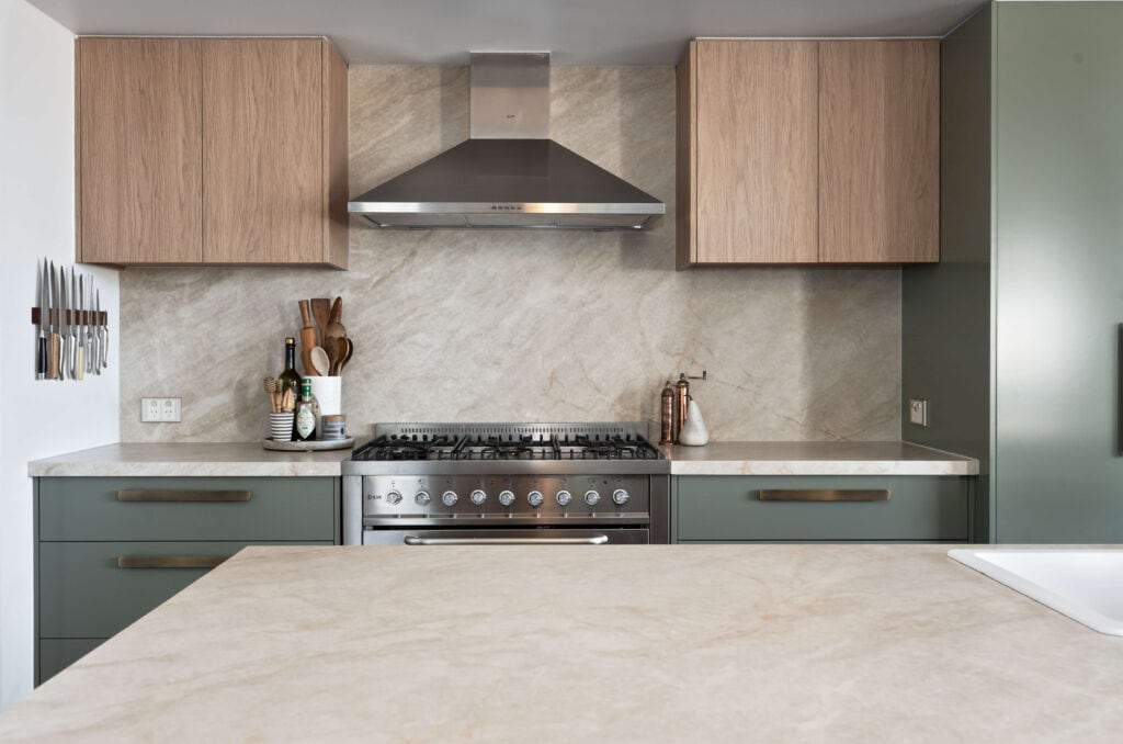 Close-up view of a modern kitchen featuring a natural stone benchtop and splashback with subtle veining, complemented by sage green cabinetry with brass handles and warm woodgrain upper cabinets. A stainless steel gas range and rangehood take centre stage, with a neatly arranged magnetic knife rack and natural timber accessories adding both style and function.