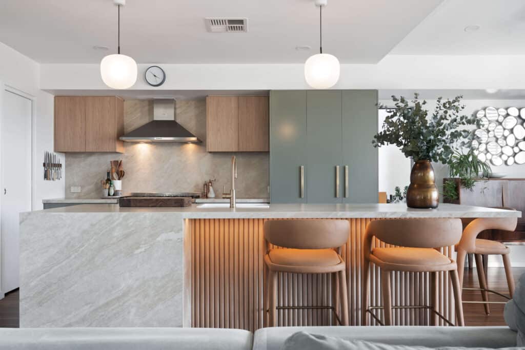 Contemporary kitchen featuring a natural stone waterfall benchtop with integrated LED lighting, fluted timber detailing on the island, and brushed brass tapware. Matte sage green cabinetry with large brass handles complements warm timber upper cabinets, while stainless steel appliances and a concealed fridge enhance the modern look. The space is accessorised with a large bronze vase filled with greenery, stylish leather bar stools, and a unique circular wall mirror, all set against timber flooring.