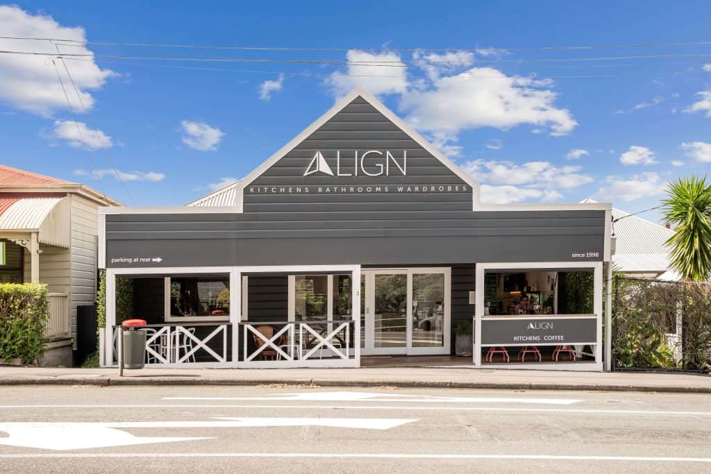 "Exterior view of the Align store specializing in kitchens, bathrooms, and wardrobes, located in a Red Hill. The building features a modern, gray facade with white accents and large front windows. A sign indicates 'Parking at rear' and another notes the business has been operating since 1998. Clear blue sky with sparse clouds can be seen above.