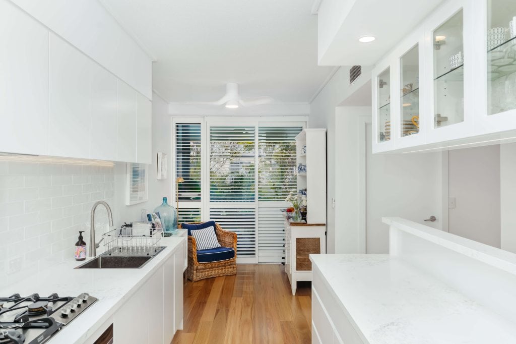 Spacious corner of a galley kitchen in a New Farm, Brisbane home, bathed in natural light from the window, featuring white cabinetry, a cozy nook with a wicker chair and decorative elements, and rich wooden flooring.