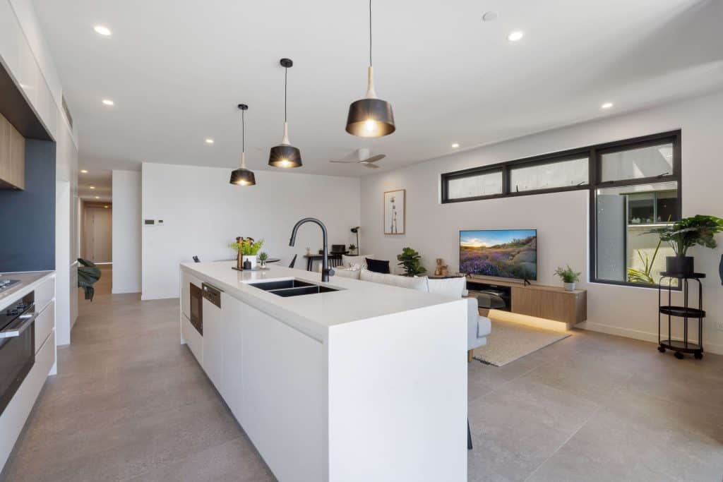 Spacious modern kitchen with an open layout flowing into the living room, featuring a large island bench, top-of-the-line appliances, and tasteful pendant lighting, complemented by greenery and a plush sofa.