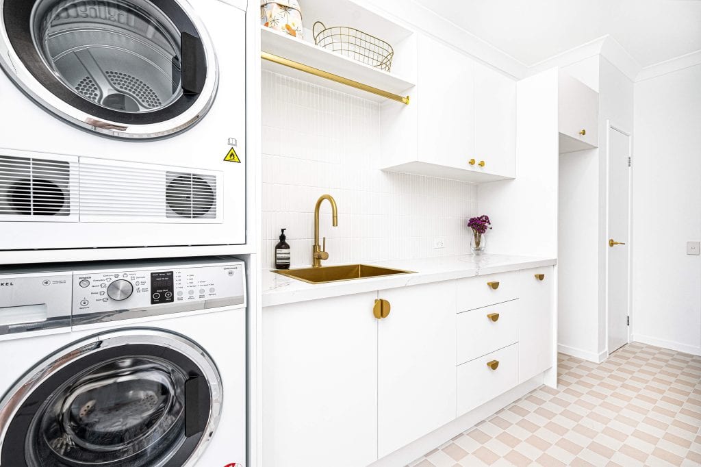 Modern laundry room featuring stacked washer and dryer with white and stainless steel finish, white cabinetry with gold handles, a built-in sink with a gold faucet, and terrazzo-style floor tiles.