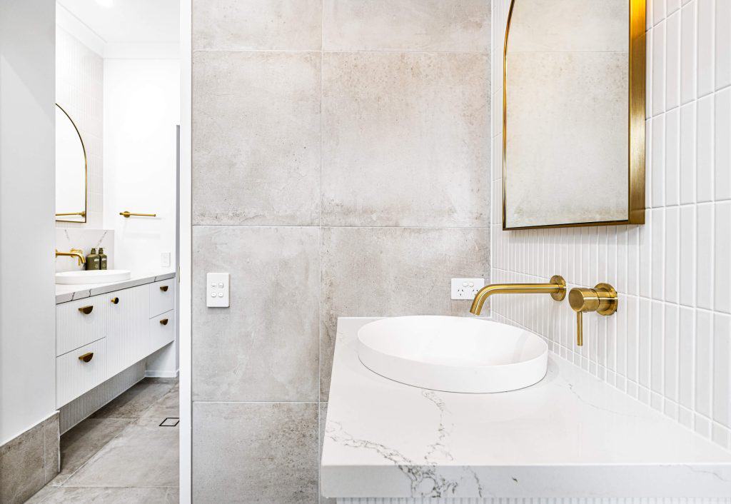 Chic bathroom detail showing a section of the vanity with a white oval vessel sink atop a marble countertop, fluted cabinet doors with gold pulls, and a rectangular mirror with a soft golden backlight, complemented by white subway tiles and large grey floor tiles.