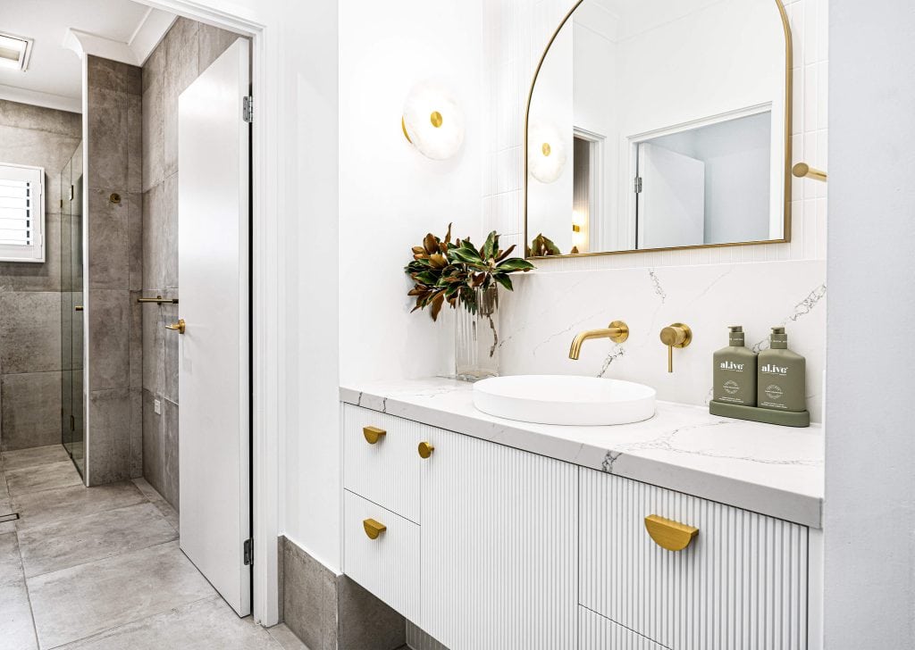 Sleek bathroom design with a side view of a white vanity, gold fixtures, a circular mirror, and a glimpse into a tiled shower area.
